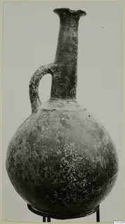 Pottery jug with a tall neck, flared rim, single loop handle and round body, standing on a metal stand.
