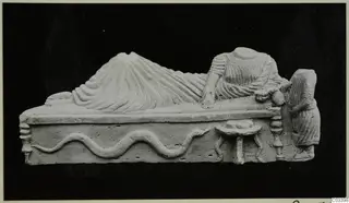 Limestone relief of a reclining draped figure on a couch, with a serpent along the base and a small table beside it