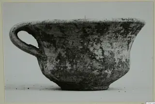 Plain white wheel-made pottery bowl with a single loop handle, seen from the side