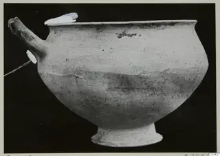 Hand-made pottery bowl with a horizontal spout-like handle on a short ring foot, photographed against a black background.