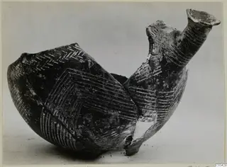 Dark pottery vessel with a tall flaring neck and incised line and chevron patterns, photographed alone