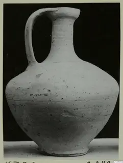 Pale ceramic jar with a single loop handle and tall neck, photographed against a black background