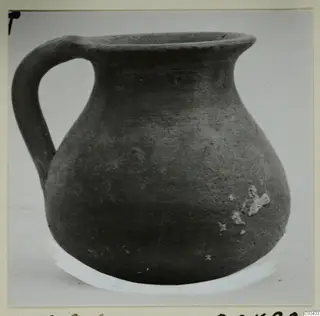 Dark grey pottery jug with a single loop handle and wide rim, photographed against a plain background.