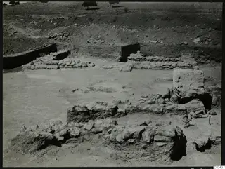 Excavation at Ayia Irini with low stone wall foundations on a levelled surface, a large squared block at right, and trees on the skyline