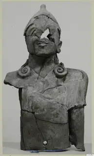 Terracotta bust of a figure with a rounded headdress, spiral shoulder ornaments and a broken face, against a plain grey background
