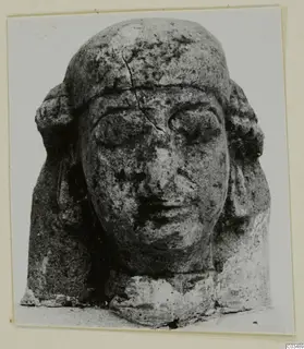 Limestone sculpted head with a flat headdress, heavy-lidded eyes and side locks, photographed on a card mount
