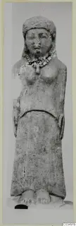 Limestone standing figure with a close-fitting head covering and necklace, arms at its sides, on a plain background