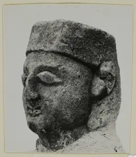 Limestone sculpture of a head wearing a flat-topped cap, with a large almond-shaped eye and a rounded ear