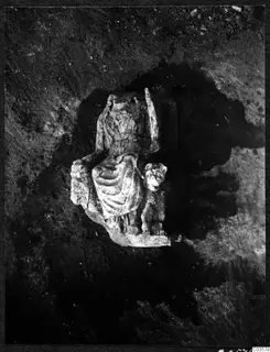 Carved stone seated figure with a smaller animal figure beside it, lying in excavated soil at Cholades, Soli