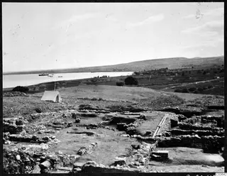 Excavated stone walls at Soli with a white tent on a mound, and a bay with boats beyond