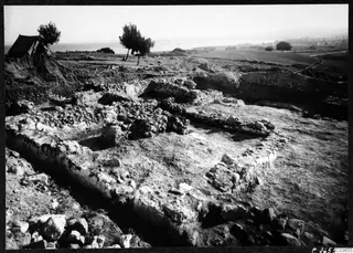 Excavated rubble stone walls of Room V at Soli, seen from the west, with a tent and trees on the left and the sea beyond.