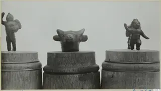 Three small bronze figurines on wooden stands: two standing human figures and an animal head with horns.