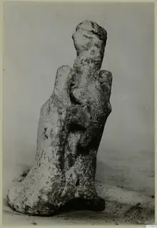 Terracotta figurine of a standing human figure with a rounded head, photographed on a grey surface
