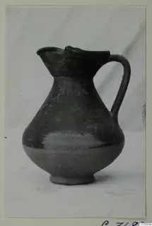Jug with a single handle and a trefoil-like pinched spout, dark surface, photographed on a plain background.