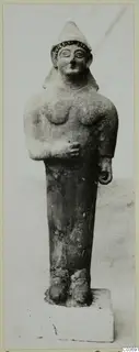 Terracotta figure with a tall conical cap and beaded headband, standing on a rectangular plinth.