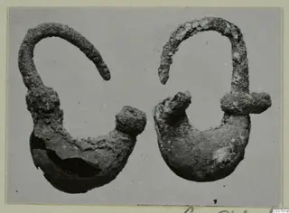 Two corroded metal objects from Vouni, each with an arched wire arm and a rounded lower body, shown on a grey ground.
