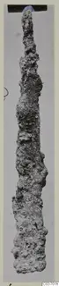 Long, narrow, heavily corroded iron tool photographed upright against a grey background, from Vouni