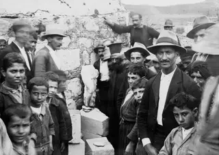 Crowd of men, women and boys gathered around a draped stone statue fragment on a block at Galini