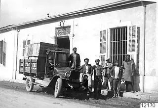 Old lorry loaded with wooden crates in front of a whitewashed building, with a group of men and a boy standing beside it.