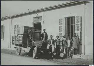 Lorry loaded with wooden crates parked outside a whitewashed building, with seven men posing beside it