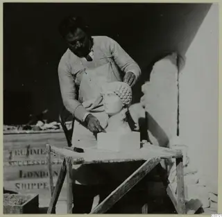 Person in an apron working on a pale sculpted female head on a wooden table, with a carved fragment behind