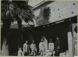 Five men stand with large clay vessels and baskets in a courtyard beside a stone building and palm trees