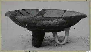 Pottery White Painted III-1 bowl on three legs, with a broken rim, standing on a dusty surface
