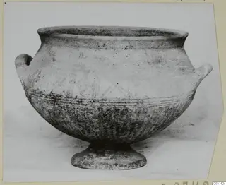 Pottery Black Slip Painted vessel with two small handles and a low foot, photographed in side view