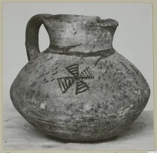 Round-bodied jug with a strap handle and dark painted bands and a cross-like motif, photographed on a plain surface.
