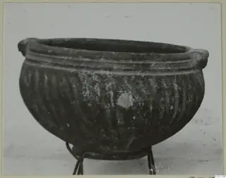 Dark-slipped bowl with vertical fluting and small rim handles, shown on a wire stand
