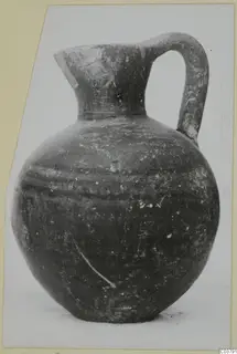 Globular jug with a single strap handle and a pinched spout, photographed and mounted on a card.