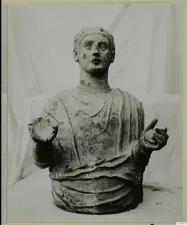 Stone bust of a draped figure with raised hands, photographed against a white cloth.
