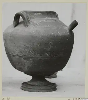Pottery vessel with a loop handle, a tubular spout and a pedestal foot, photographed from the side.