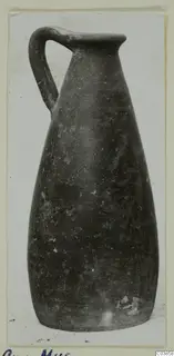 Tall dark jug with a single loop handle and a flared rim, photographed on a card mount