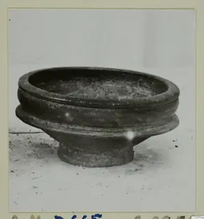 Dark pottery bowl with a wide rim and a stepped body on a short foot, photographed on a pale surface.