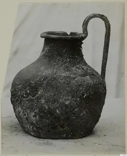 Corroded bronze pot with a rounded body, flared mouth and a single tall strap handle.