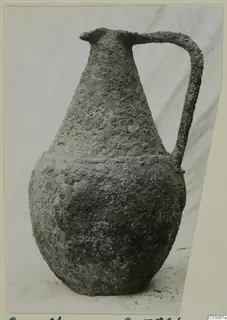 Corroded bronze vessel with a long neck and a single strap handle, shown against a light cloth backdrop