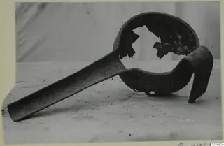 Corroded metal ladle with a long flat handle and a broken bowl, shown on a light surface.