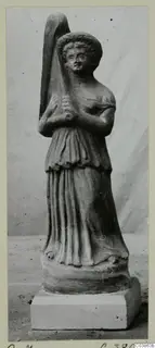 Terracotta figurine of a standing female figure with curly hair, holding a tall object, on a rectangular plinth
