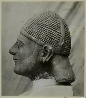 Limestone head in profile, wearing a cap covered in rows of raised knobs, on a plain base.