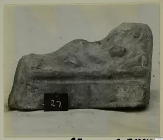 Grey fragment of a worn terracotta or stone object with raised forms, shown with a number card at its base