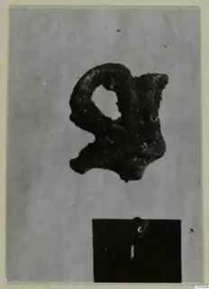 Corroded bronze fragment with a loop and a hole, photographed beside a number label, from Amathus