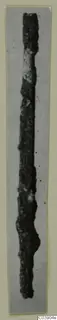 Long, thin, corroded bronze pin-like object, shown upright against a grey background.