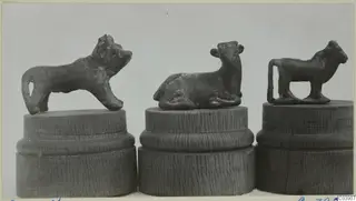 Three small bronze animal figurines, each on a turned wooden stand, photographed side by side against a grey backdrop