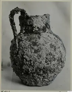 Corroded bronze jug with a looped handle, photographed against a plain grey background.