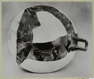 Shallow painted pottery bowl with a single loop handle, seen from above, with pale figure outlines and white restored areas.