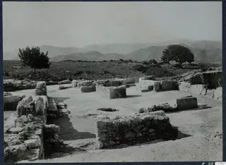 Ruined stone walls and a round hole in a flat paved area at Vouni palace, with trees and hills behind.