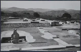 Stone steps and low wall foundations of the palace ruins at Vouni, with a stone figure in the foreground and hills behind.