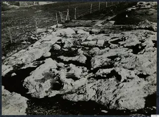 Stone foundations and cut bedrock at Vouni, with a wire fence and a stone building beyond.
