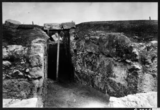 Eisodos crypt at Soli: a narrow passage between stone walls, with a timber frame over a dark opening and excavated ground in front.